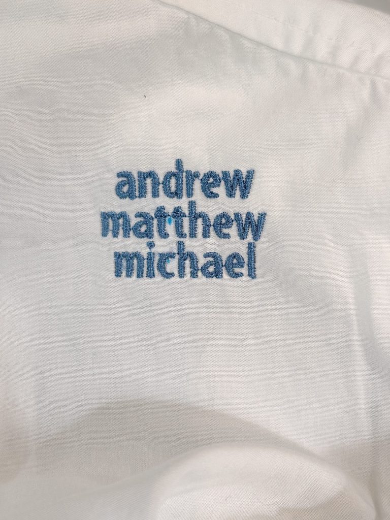 White fabric with blue embroidered text reading "andrew matthew michael" in lowercase letters, arranged in three lines.