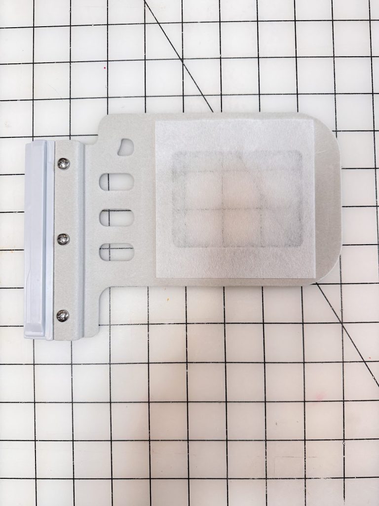 A metal embroidery hoop with a white stabilizer sheet is placed on a grid-patterned cutting mat.
