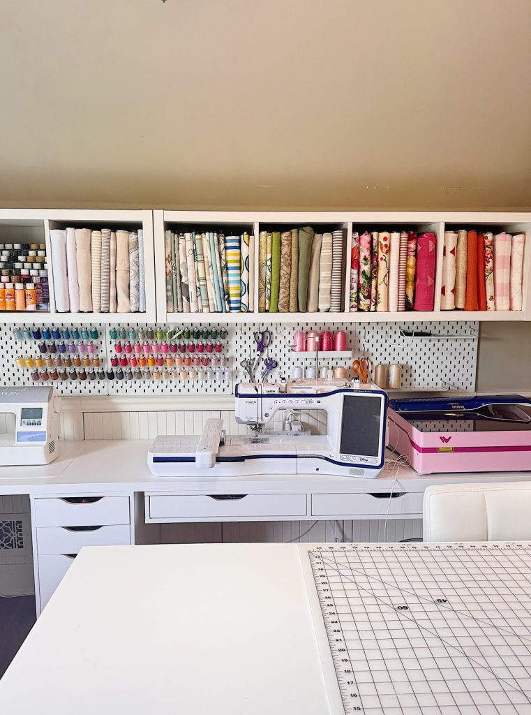 A sewing room with a sewing machine, organized fabric rolls, threads, craft supplies on shelves, and a cutting mat on a white table.