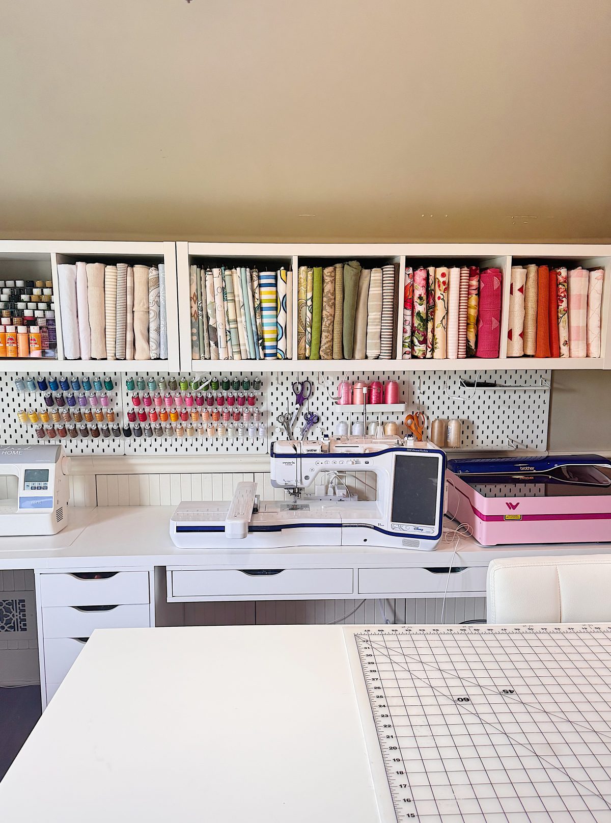A sewing room with a sewing machine, organized fabric rolls, threads, craft supplies on shelves, and a cutting mat on a white table.