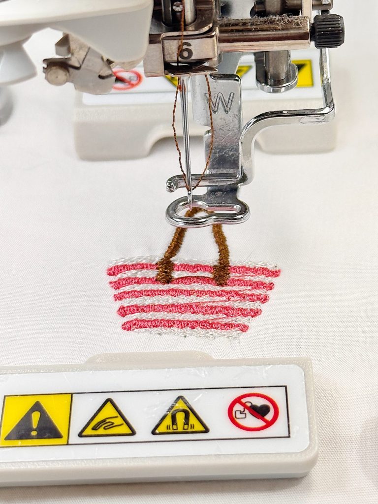 Close-up of a sewing machine stitching red and brown threads onto white fabric, with safety warning symbols visible on a white panel.