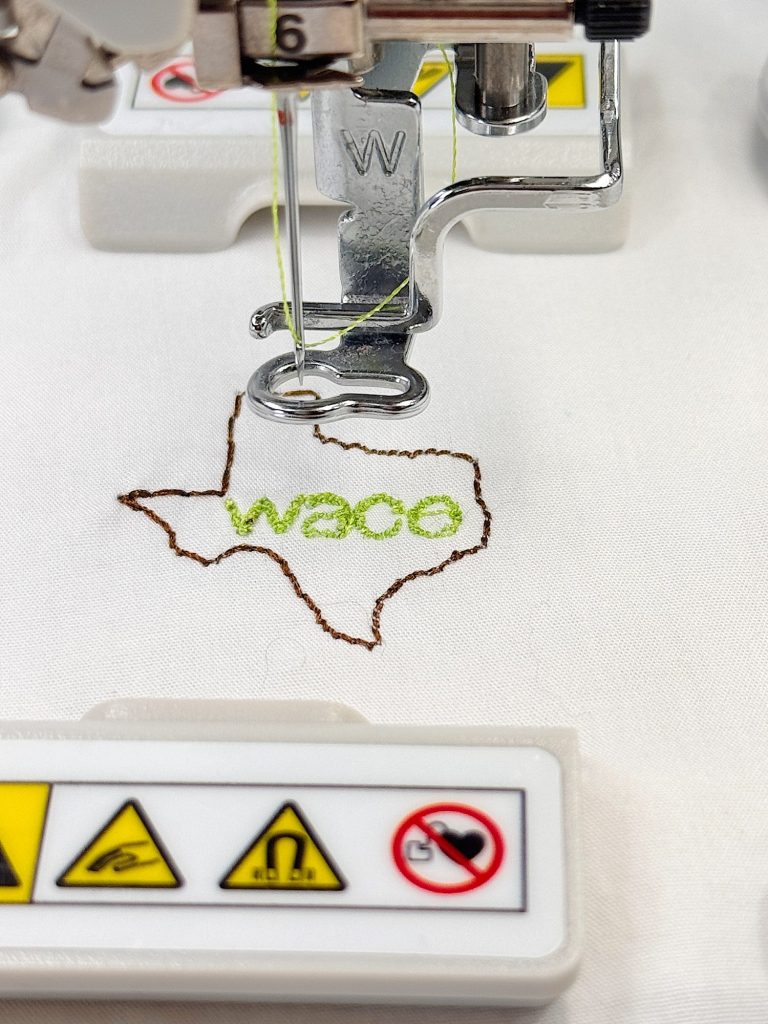 A sewing machine stitches an outline of Texas with the word "Waco" inside on white fabric; safety warning labels are visible in the foreground.