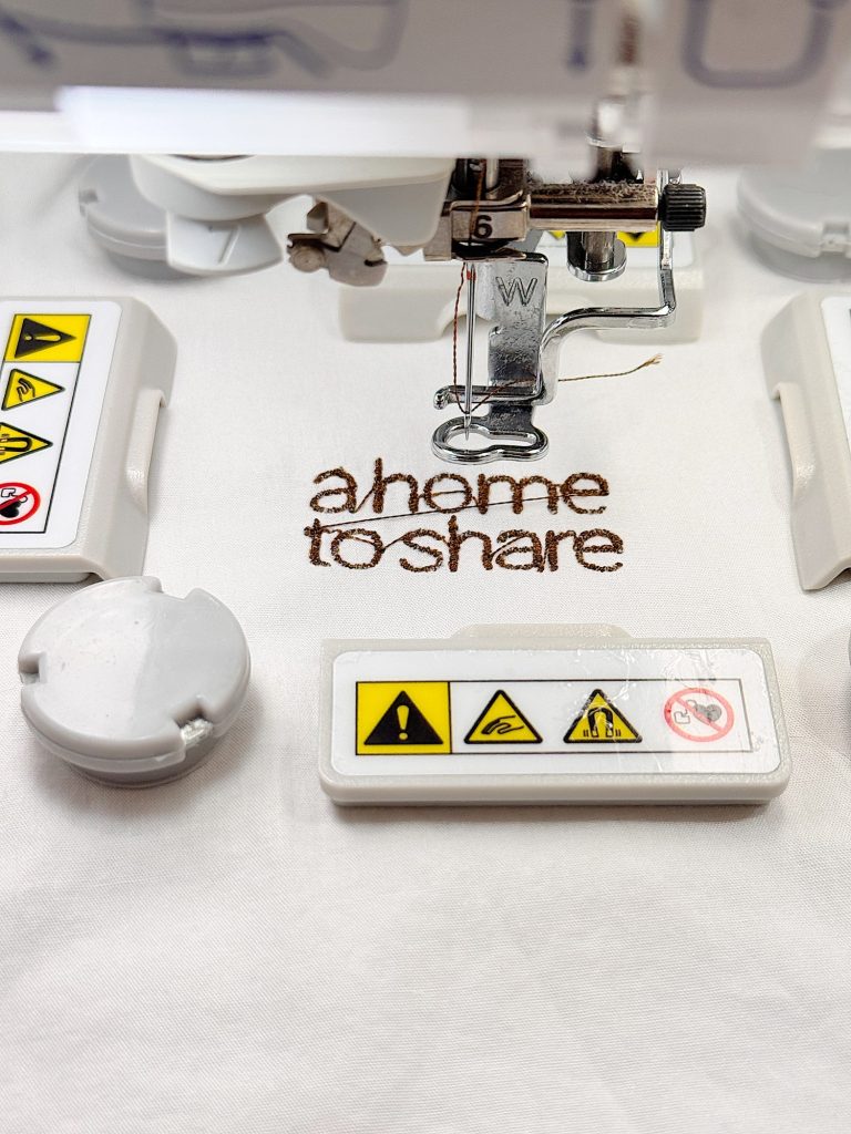 Close-up of an embroidery machine stitching the words "a home to share" on white fabric, surrounded by warning label components.