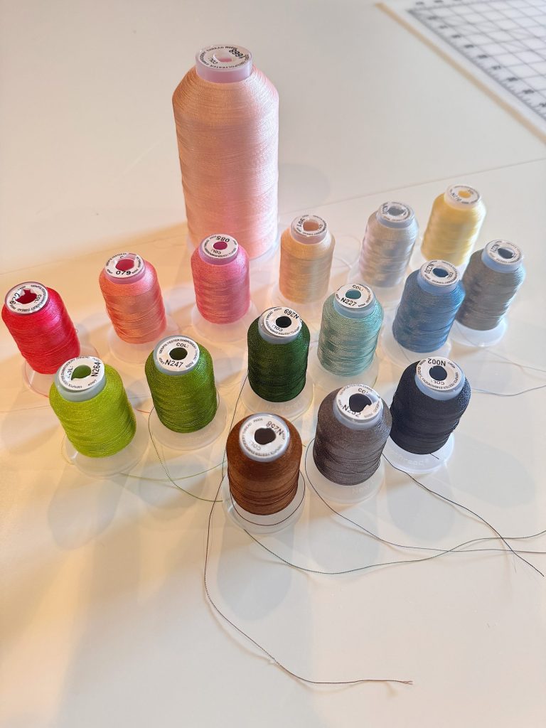 Spools of thread in various colors, including pink, red, green, brown, black, and yellow, are arranged on a white surface next to a ruler.