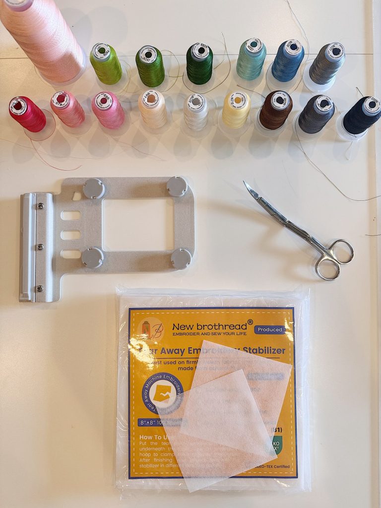 Spools of colored thread, embroidery scissors, a metal sewing bracket, and a pack of tear-away embroidery stabilizer are arranged on a light surface.