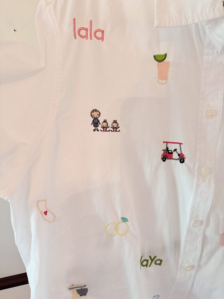 White shirt with colorful embroidered designs including text, a small family, a drink, a golf cart, California outline, wedding rings, and the word "yay.