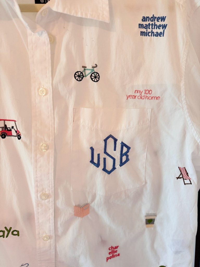 White button-up shirt with various embroidered designs including text, a bicycle, golf cart, book, chair, and initials "USB" on the pocket.