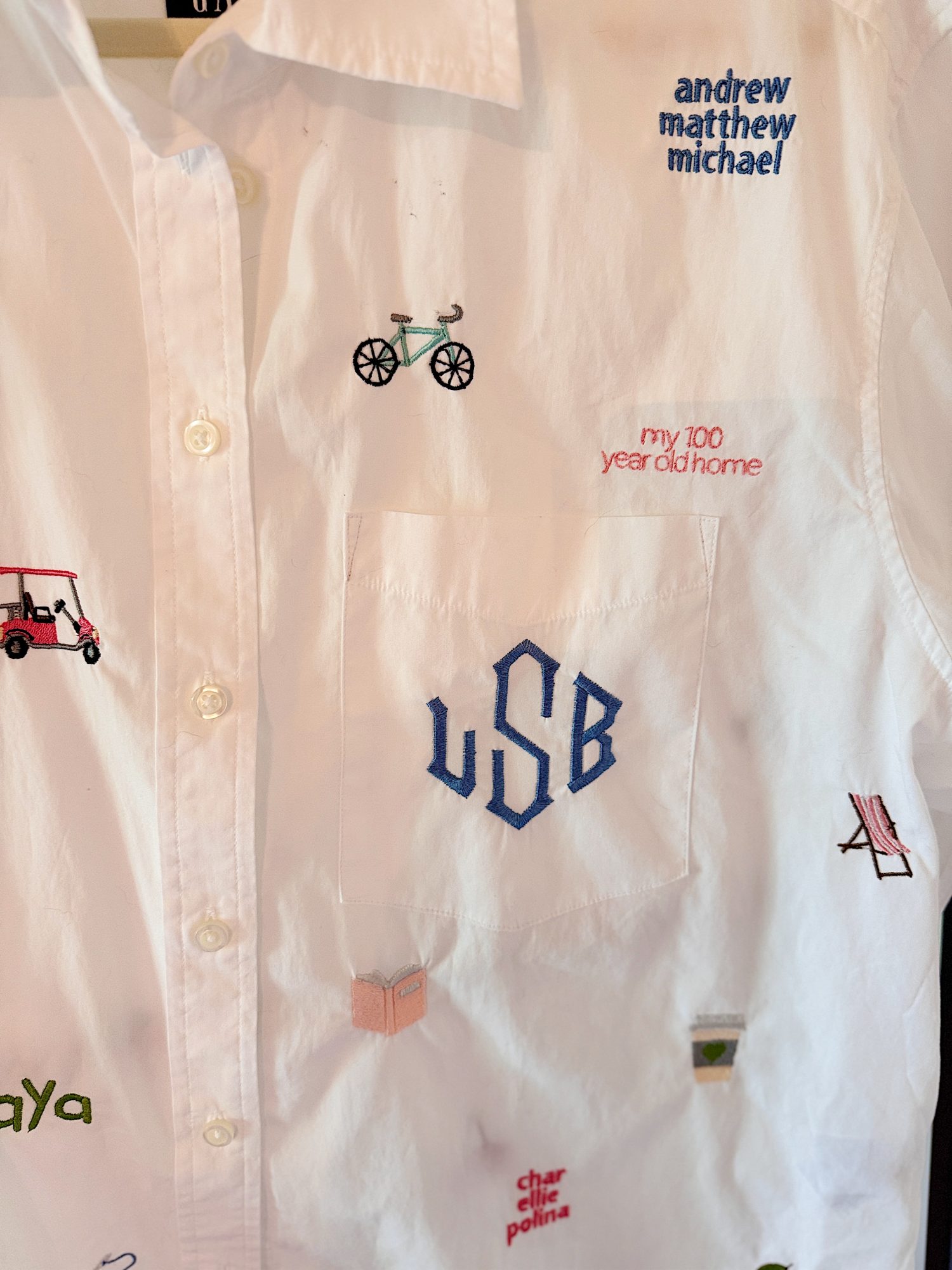 White button-up shirt with various embroidered designs including text, a bicycle, golf cart, book, chair, and initials "USB" on the pocket.