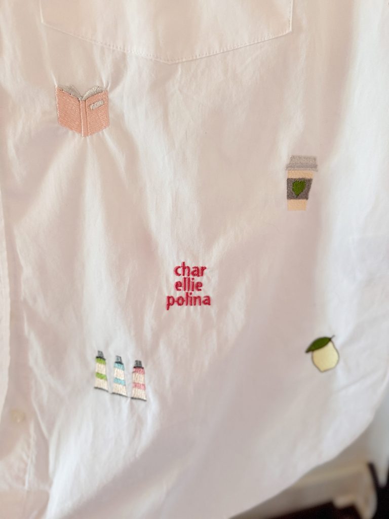 White fabric with embroidered designs including a book, a coffee cup, bottles, a lemon, and the words "char ellie polina" stitched in red thread.