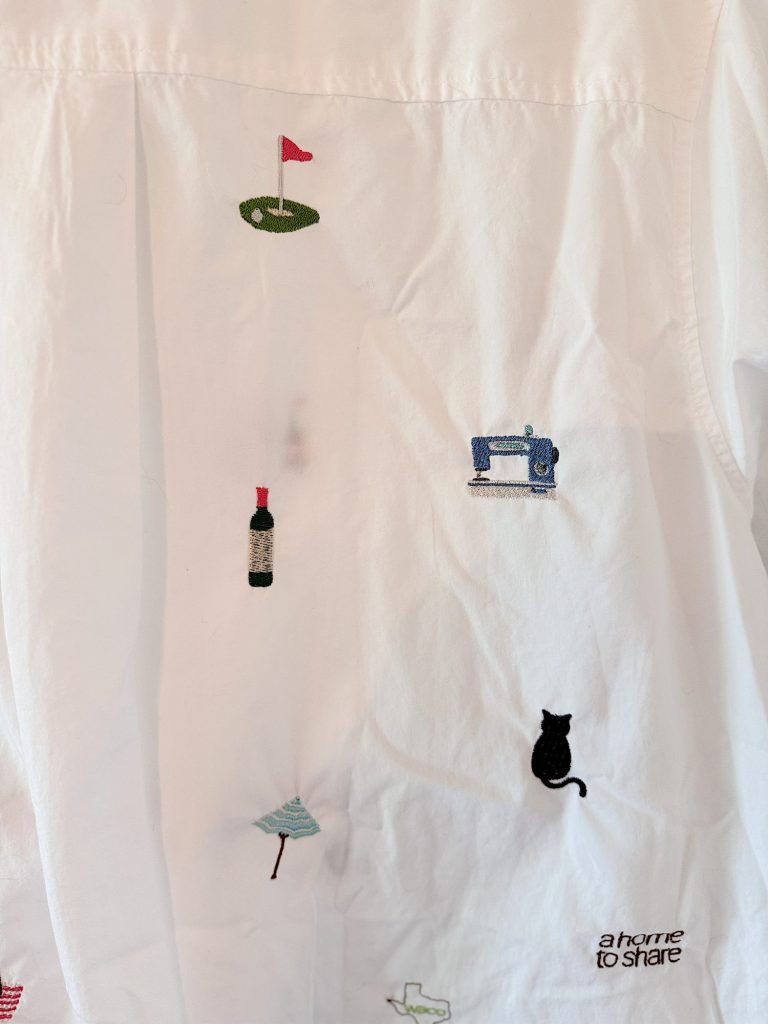 White fabric with embroidered images of a golf hole, sewing machine, wine bottle, broom, black cat, and text "a home to share.