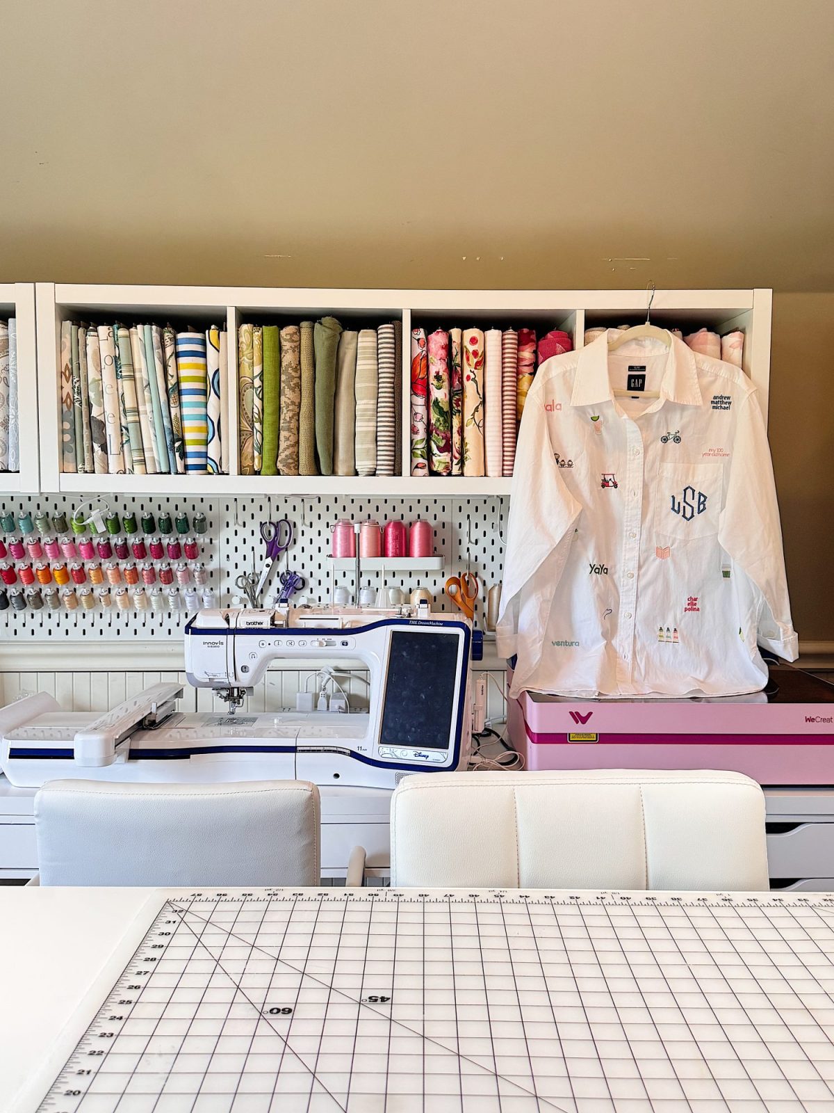 A sewing workspace with a sewing machine, threads, fabric rolls, and a white jacket displayed on a pink box, with a grid cutting mat on a table in the foreground.