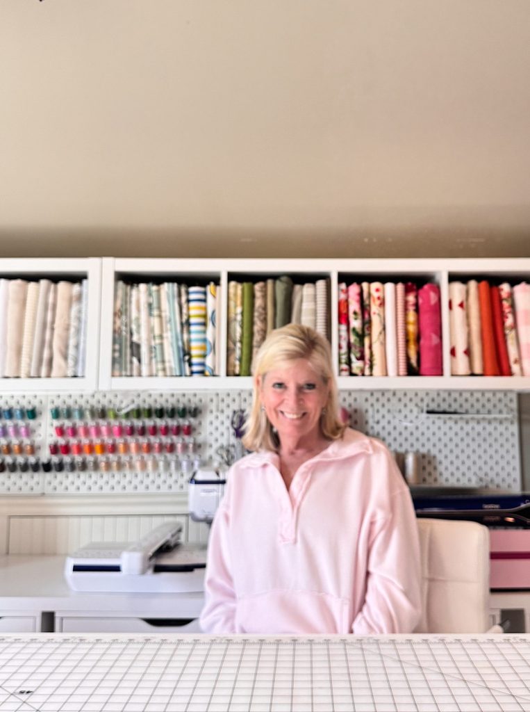 A woman in a pink sweatshirt sits at a table in a sewing room with fabric and thread neatly organized on shelves behind her.