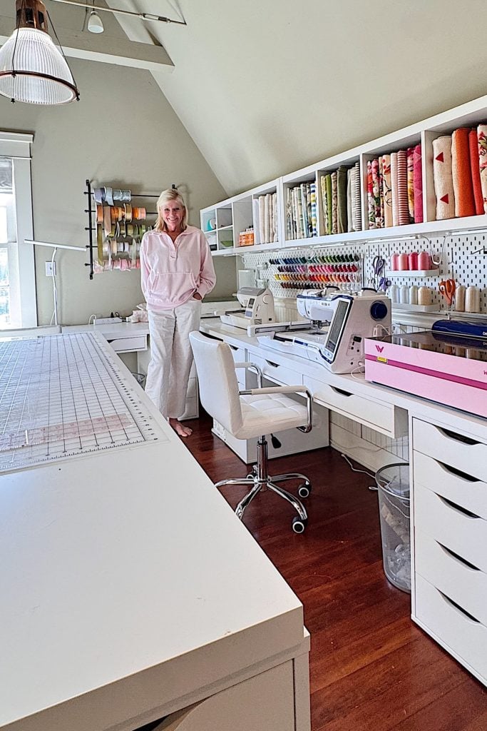 A woman stands in a well-organized sewing room with shelves of fabric, thread, and two sewing machines on a white desk.