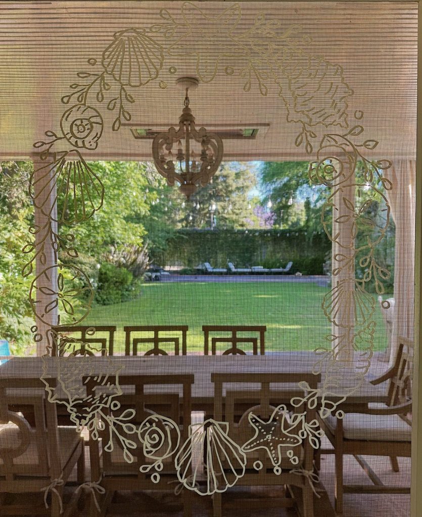A view of a backyard garden with a wooden dining table and chairs on a patio, seen through a door adorned with sea-themed decorations on the glass and charming window wreaths.