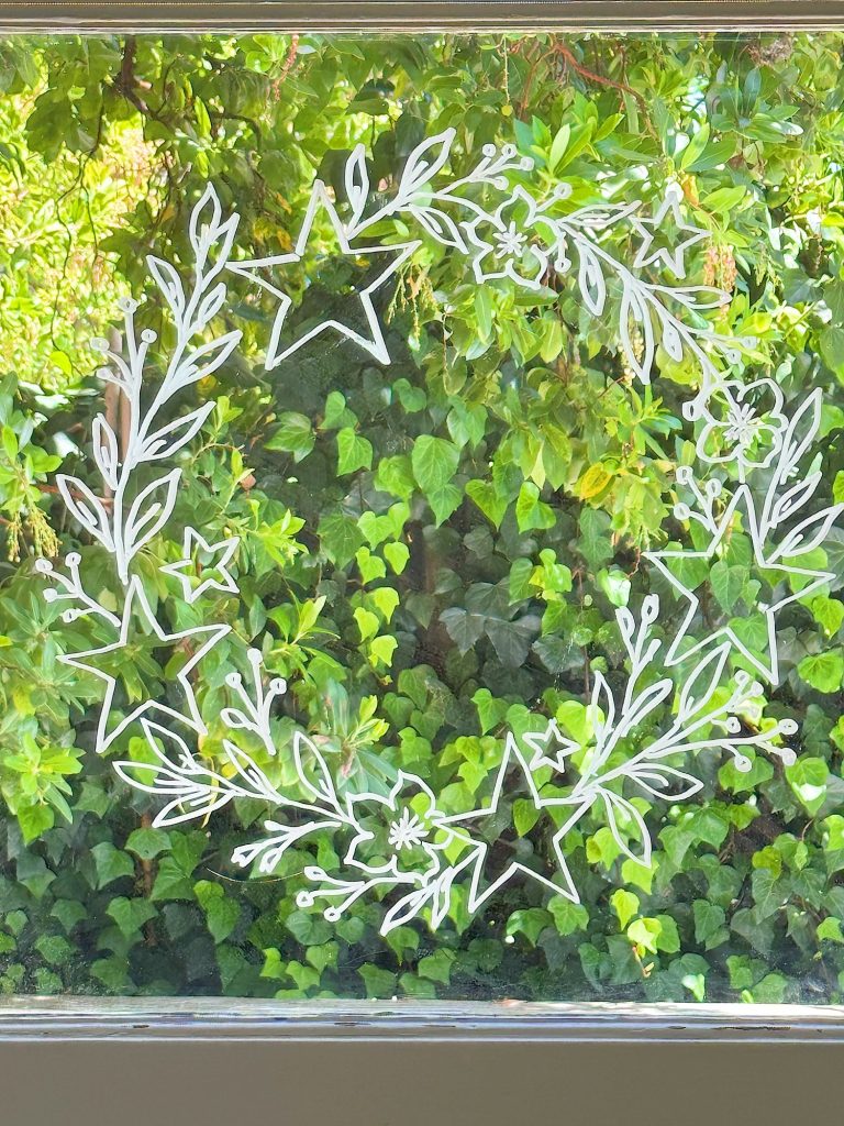White wreath and star designs are drawn on a glass window. Green leafy plants and trees are visible outside through the glass.