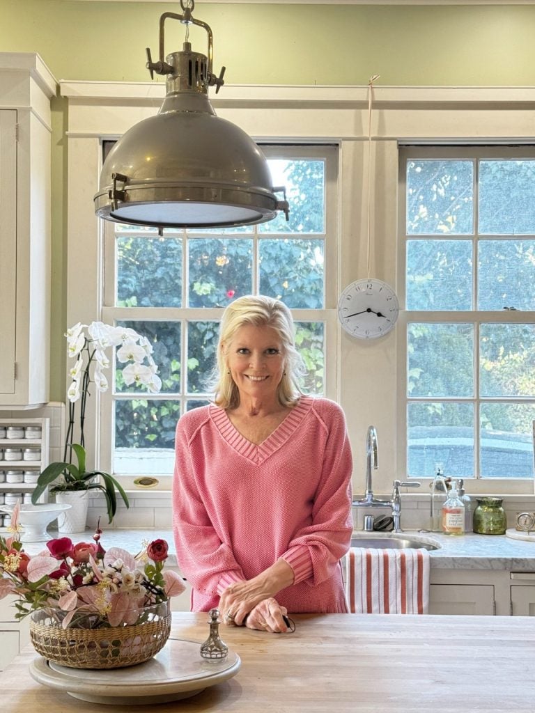 A woman in a pink sweater stands at a kitchen counter with a flower arrangement, in front of large windows with a clock on the wall.