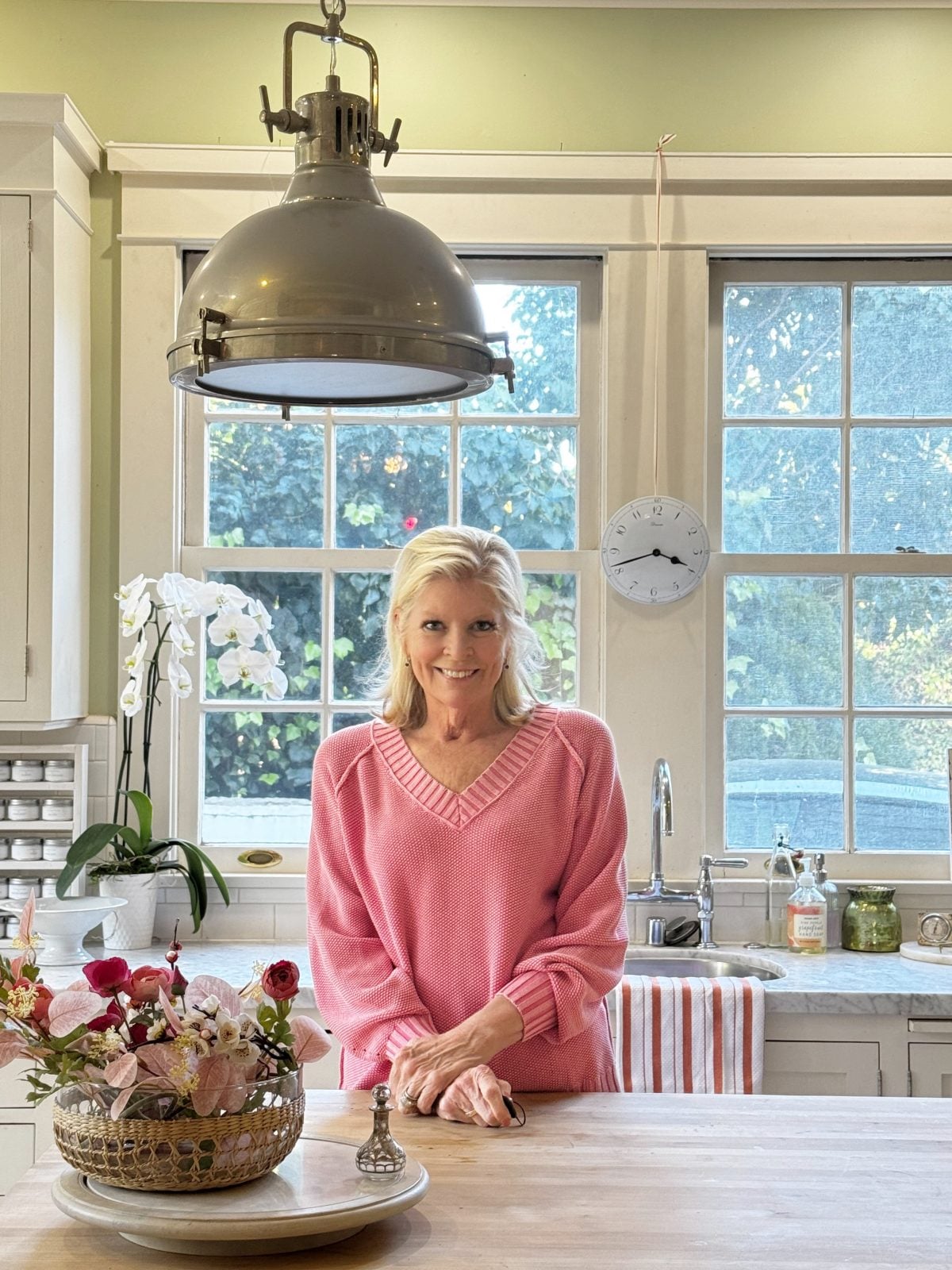 A woman in a pink sweater stands at a kitchen counter with a flower arrangement, in front of large windows with a clock on the wall.