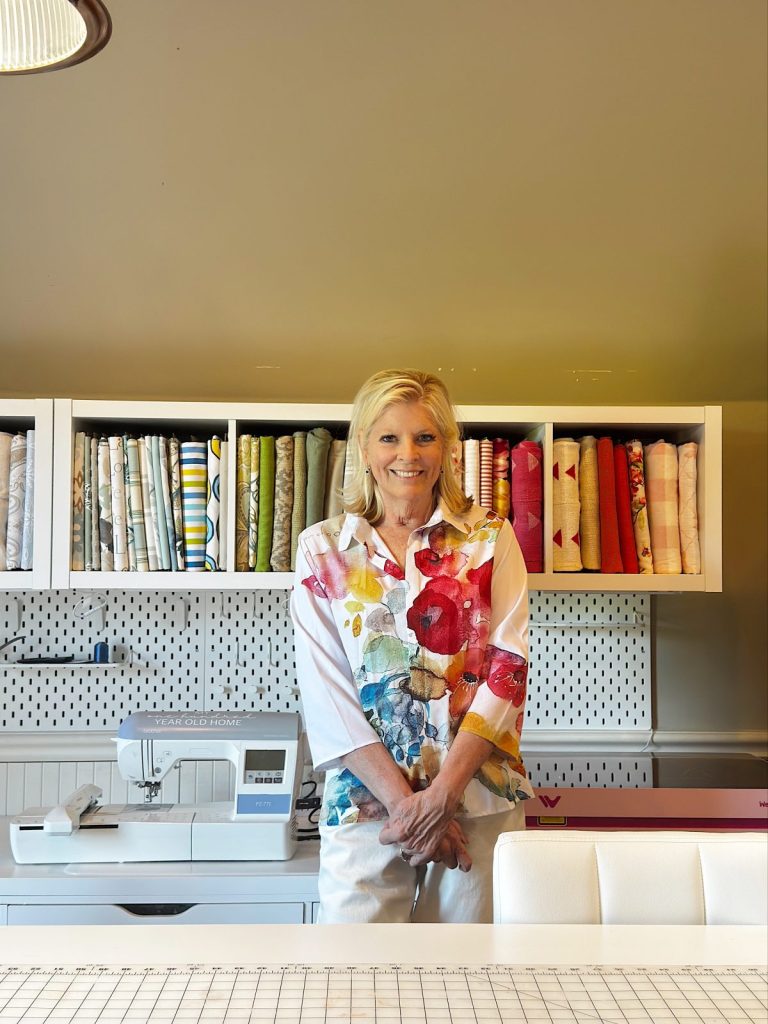 A woman stands in front of shelves filled with colorful fabric rolls, next to a sewing machine in a well-organized craft room.