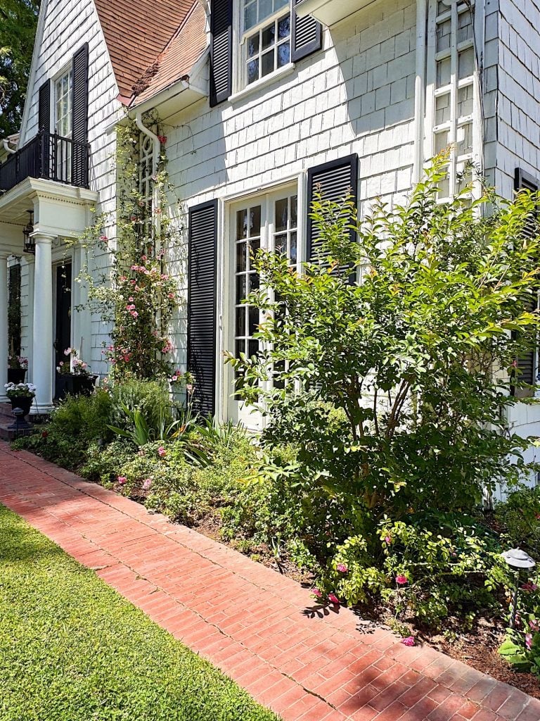 White house with black shutters, surrounded by a garden with flowering plants and shrubs, adjacent to a red brick walkway and green lawn.