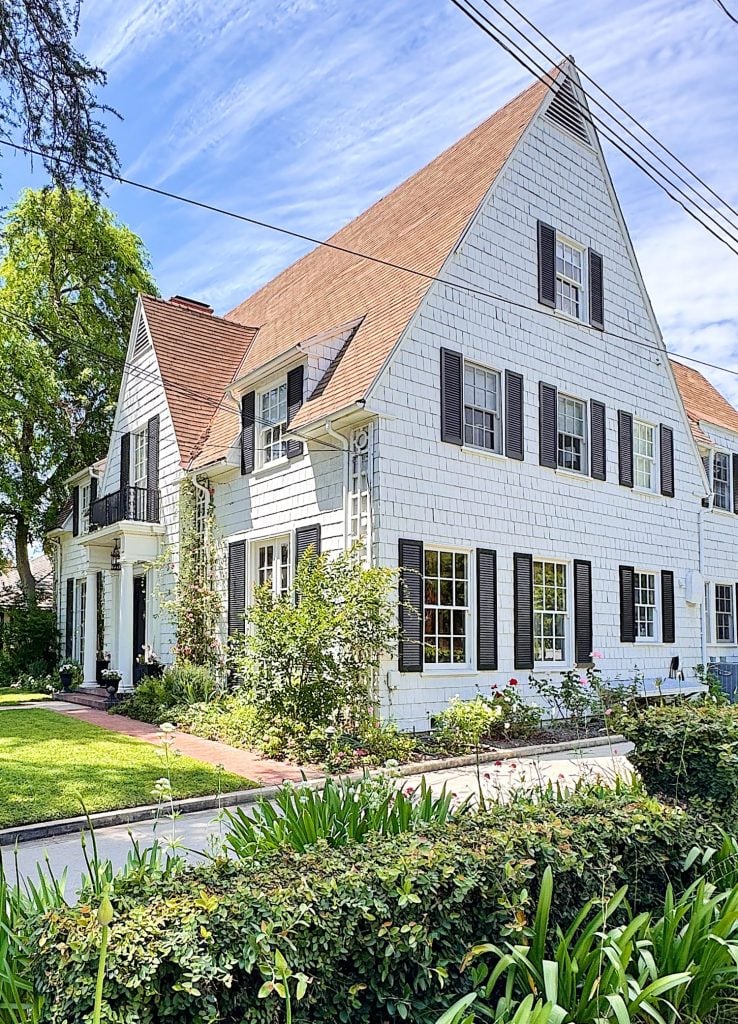 A large white two-story house with a steep gabled roof, black shutters, and a well-kept garden in front.