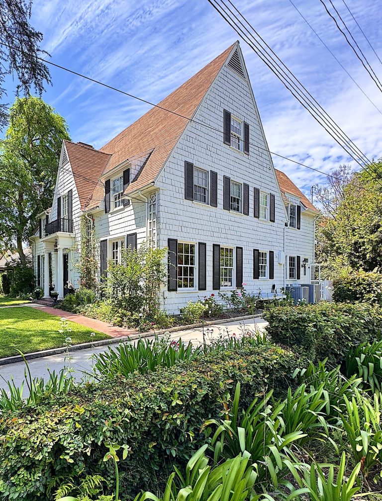 A two-story white house with brown roof shingles, black shutters, and a front yard with green plants and shrubs on a sunny day.