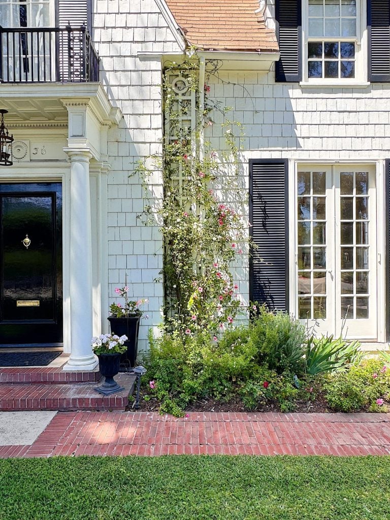 Front view of a house with white shingles, black door, large windows with shutters, and a garden with climbing plants and flowers along a brick path.