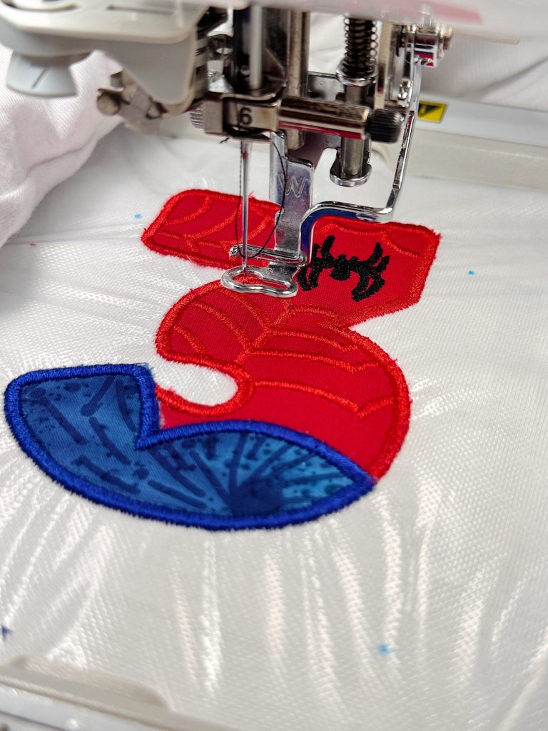 A sewing machine is embroidering a red and blue number 3 with a black spider design onto white fabric.