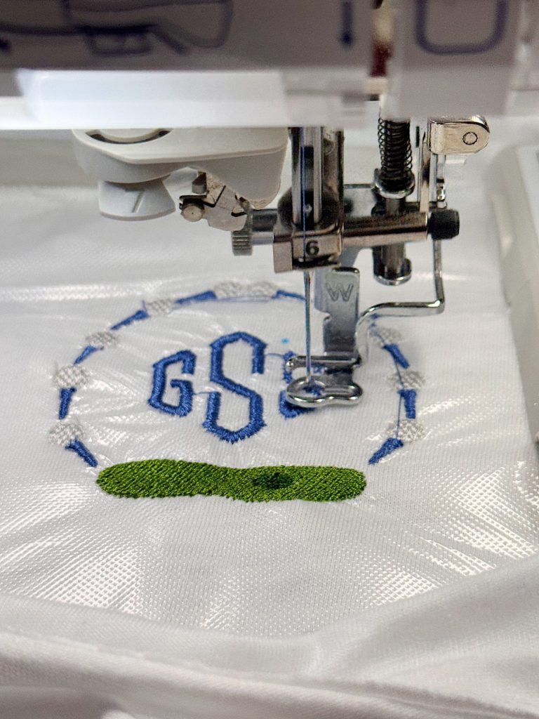 A close-up of an embroidery machine stitching the letters "GS" in blue and a green shape onto white fabric.