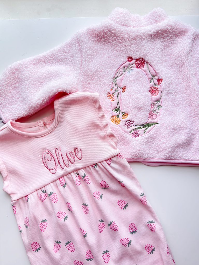 Two pink baby clothes: a fleece jacket with a floral letter "O" and a dress with "Olive" embroidered and a strawberry pattern.