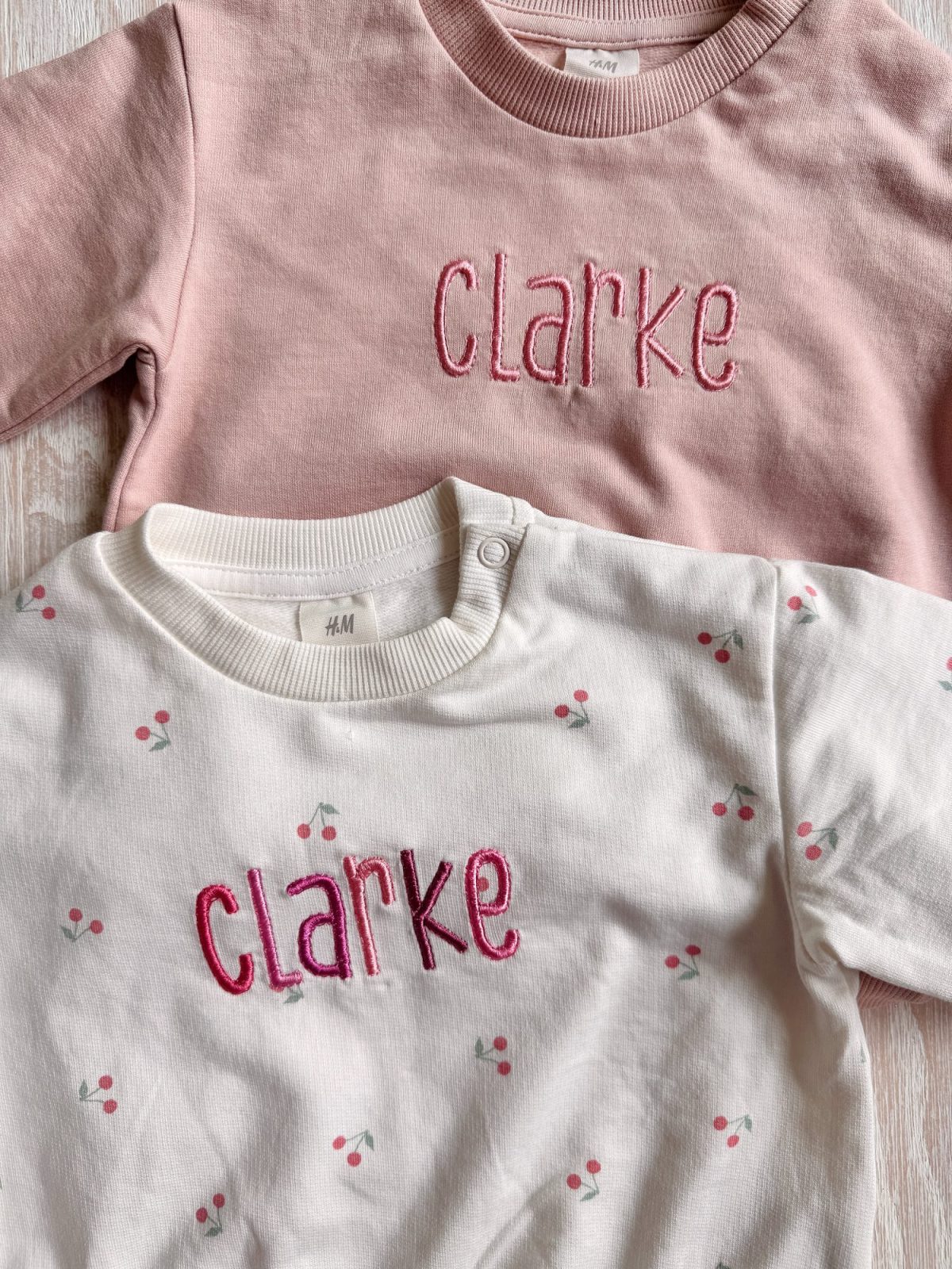 Two children's sweaters, one pink and one white with small cherry patterns, both embroidered with the name "clarke" in pink lettering.