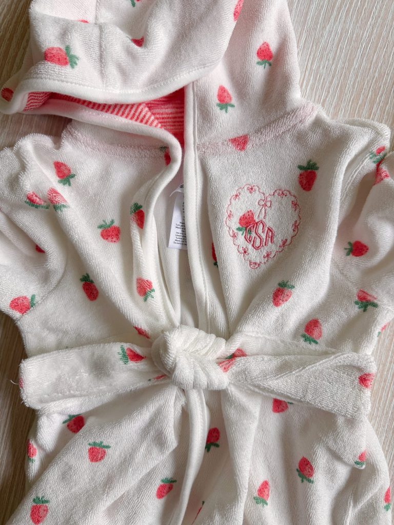 A white hooded baby robe with a strawberry pattern, a tie belt, and a heart-shaped embroidery with "USA" on the chest, laid flat on a light wooden surface.