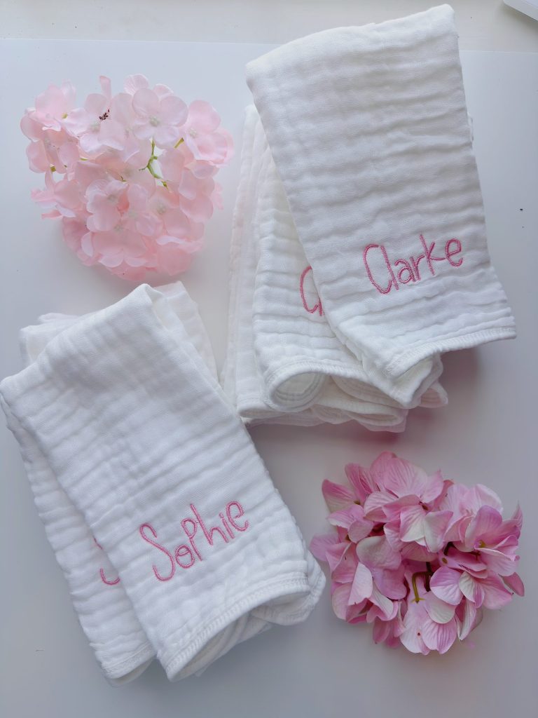 Three white muslin cloths with pink embroidered names "Sophie" and "Clarke," placed beside pink hydrangea flowers on a white surface.
