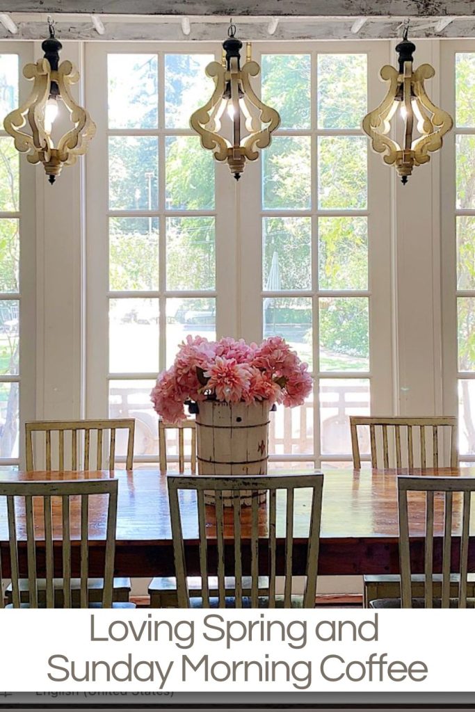 A wooden dining table with a bucket of pink flowers, surrounded by chairs, is set in front of large windows; three ornate lights hang above. Text reads: "Loving Spring and Sunday Morning Coffee.