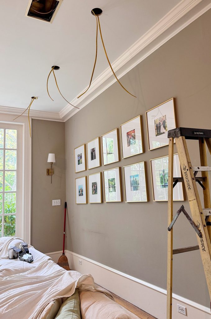 A ladder stands in a bedroom with tools on the bed, broom on the floor, and electrical wires hanging from the ceiling above framed photos on the wall.