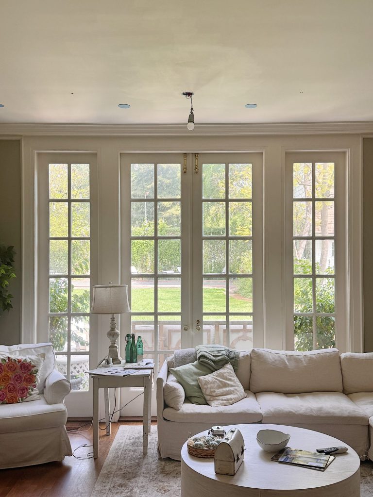 A bright living room with large windows, two white sofas, a side table with a lamp and bottles, and a coffee table with books and a bowl, overlooking a green garden outside.