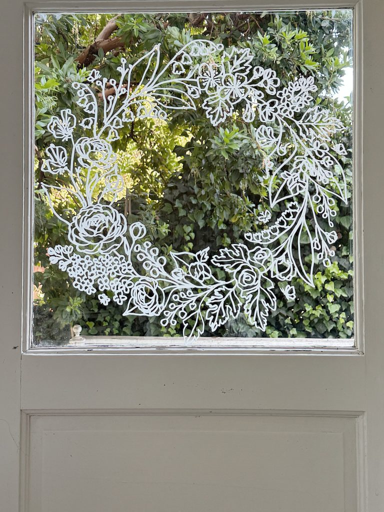 White botanical wreath design is drawn on a window pane, with green foliage visible outside through the glass.