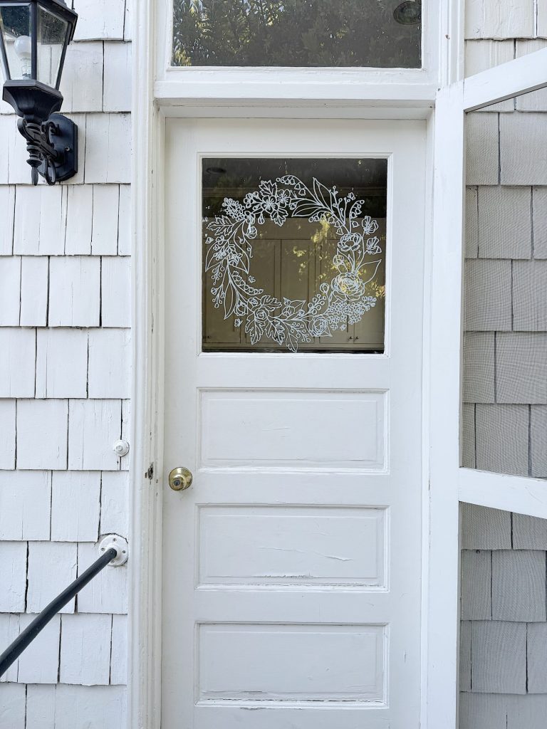 White door with a glass upper panel featuring a decorative white floral wreath design; outdoor lantern and gray shingle siding are visible.
