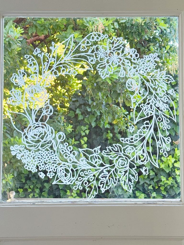 A white floral and leafy wreath is drawn on a glass window, with green foliage visible outside in the background.