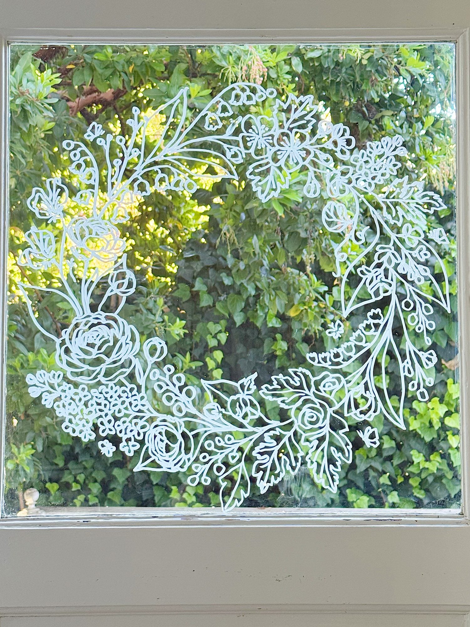 A white floral and leafy wreath is drawn on a glass window, with green foliage visible outside in the background.