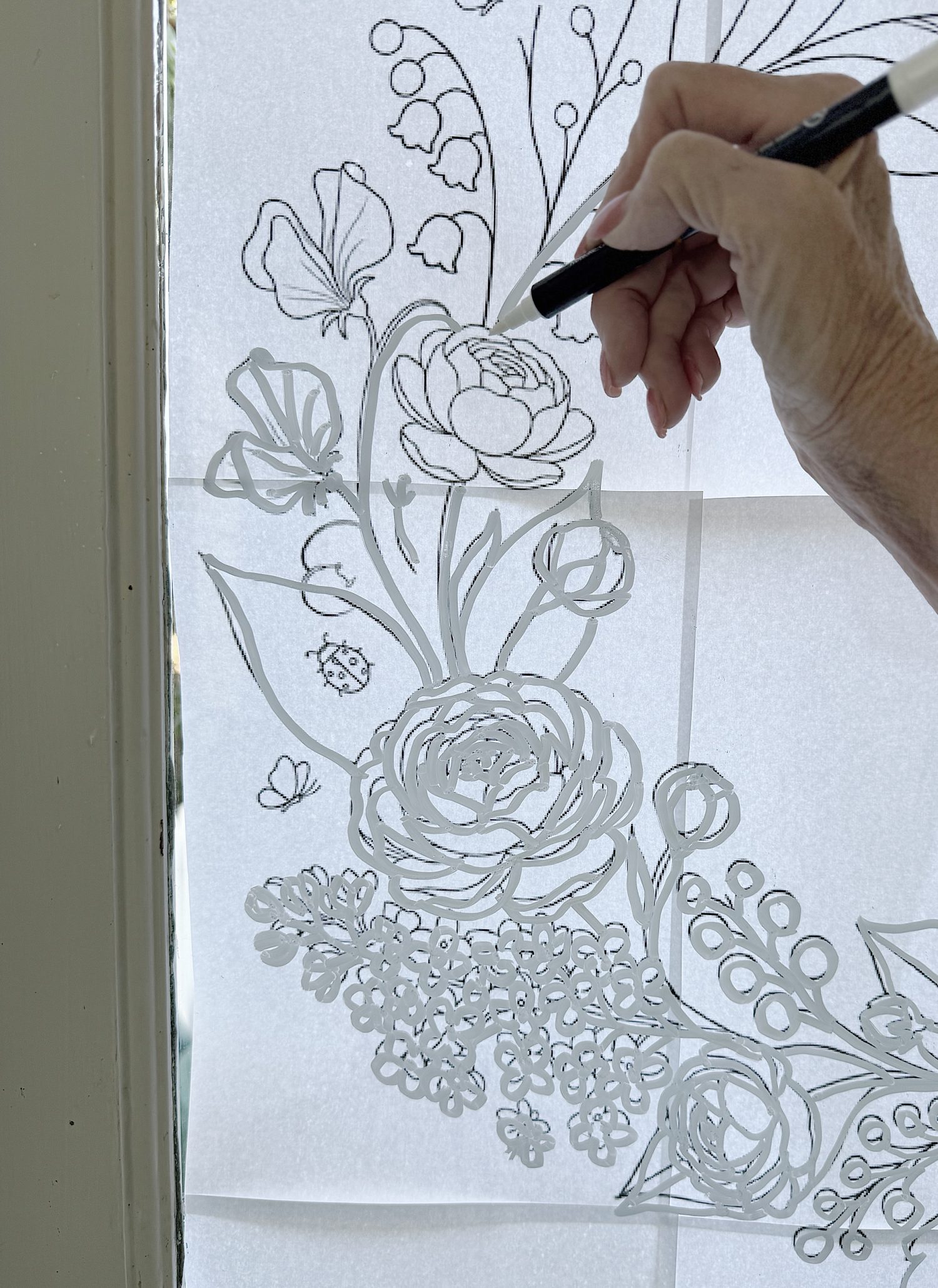 A hand holding a pen traces a floral design onto a sheet of paper taped to a window, using natural light for visibility.