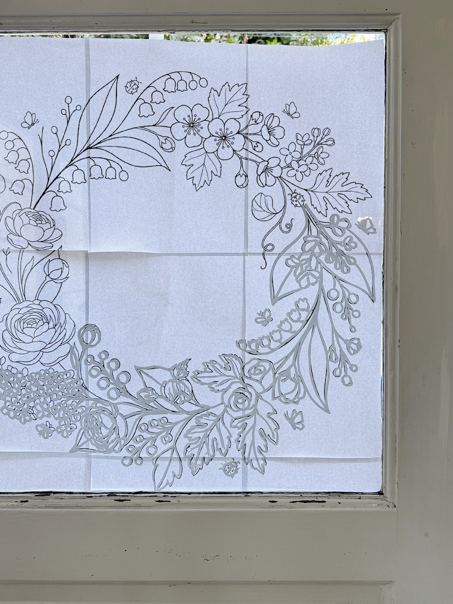 A floral wreath outline is taped to a window, featuring roses, leaves, berries, and small flowers, all drawn in black on white paper.