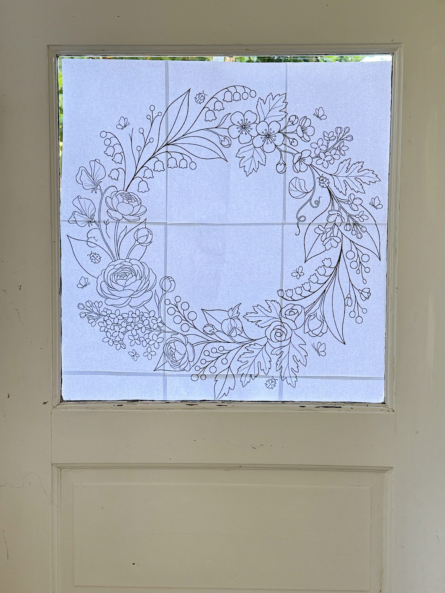 A square window with a paper taped to it, displaying a floral wreath outline drawing in black ink.
