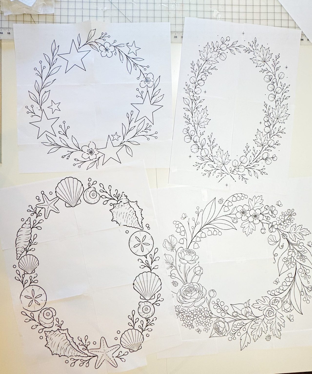 Four sheets of paper display hand-drawn circular wreath designs featuring stars, flowers, seashells, and berries, placed on a white surface.