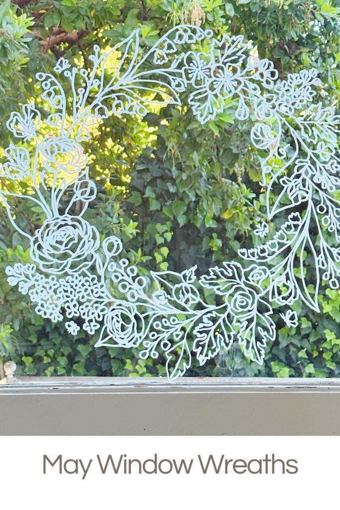 A white floral wreath design is drawn on a window with leafy greenery visible outside. Text below reads, "May Window Wreaths.