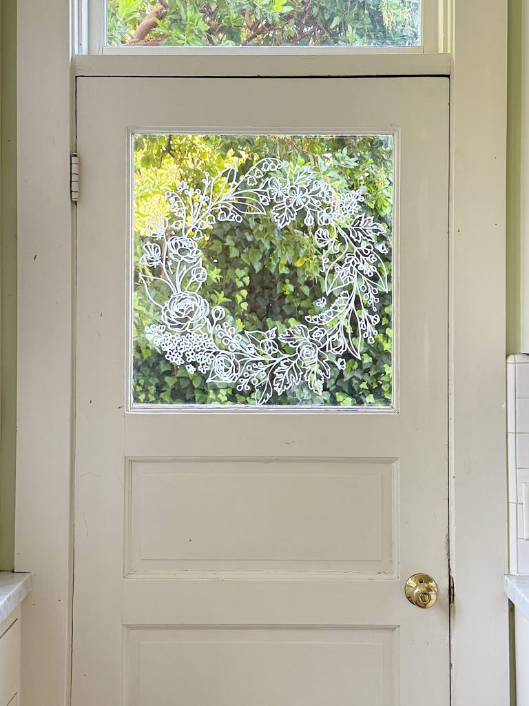 A white door with a glass window features a detailed white floral wreath design drawn on the glass, with greenery visible outside.