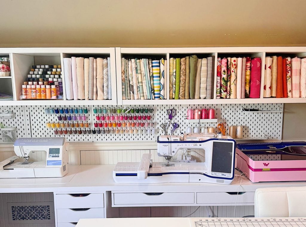 A well-organized sewing room with two sewing machines, shelves of fabric, thread spools, and craft supplies neatly arranged on a white desk and pegboard.
