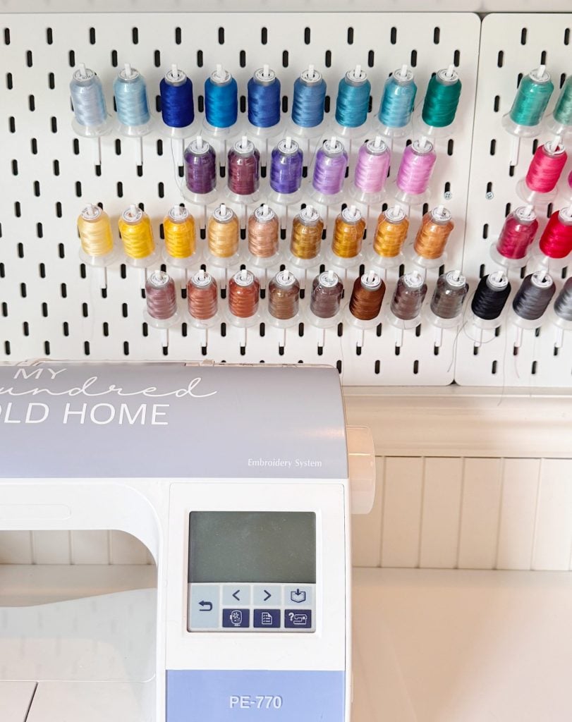 A pegboard with colorful spools of thread is mounted above an embroidery machine with a digital display and control buttons.