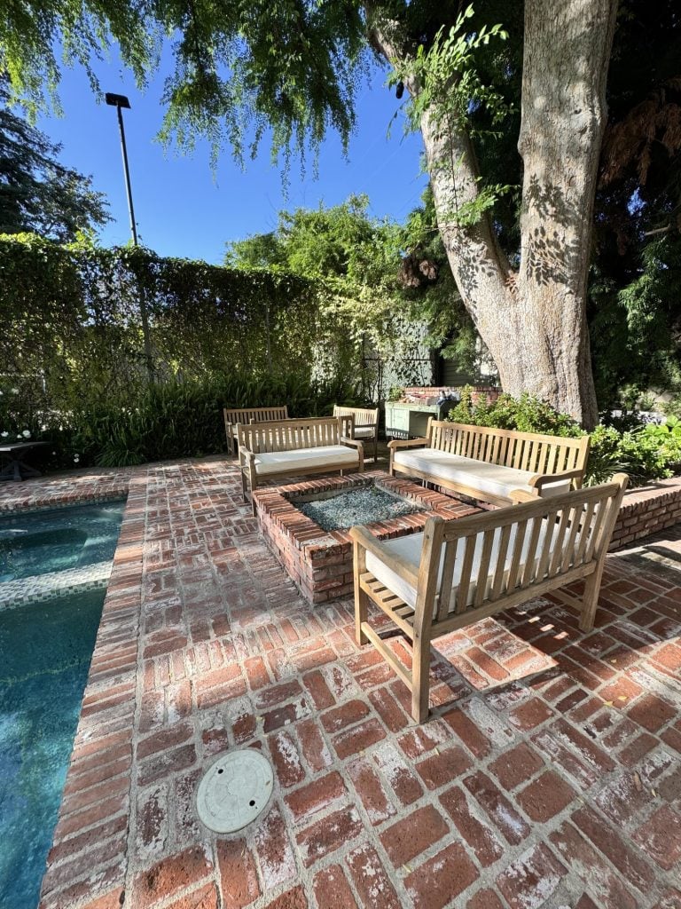 Wooden benches surround a glass-top fire pit on a brick patio next to a pool, shaded by large trees in a garden setting.