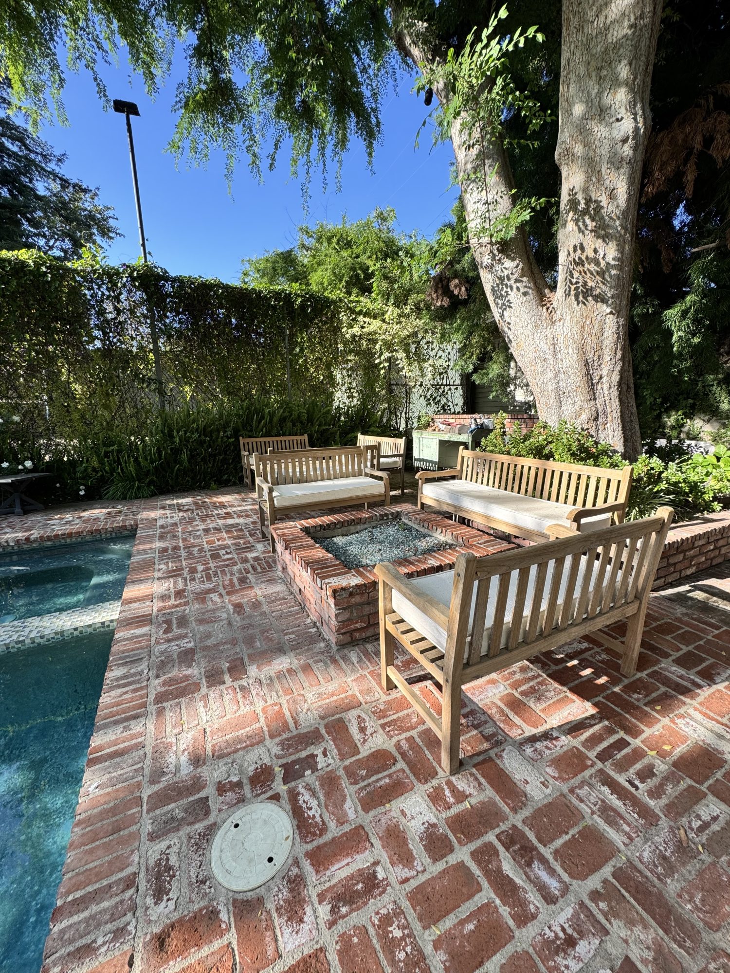 Wooden benches surround a glass-top fire pit on a brick patio next to a pool, shaded by large trees in a garden setting.