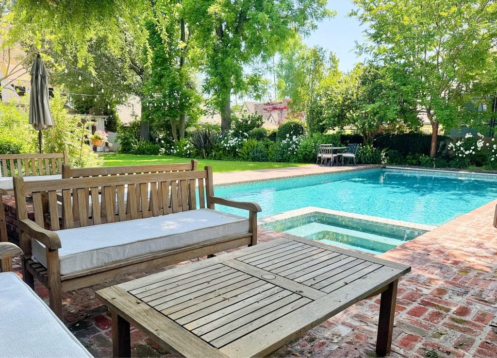 Wooden patio furniture sits on a brick patio beside a rectangular swimming pool and hot tub, surrounded by green trees and a grassy yard on a sunny day.